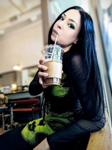 Dark-haired woman in green and black outfit, sipping iced coffee at cafe, expression of calm defiance